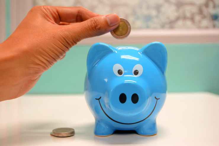 A hand placing a coin into a blue piggy bank with a smiling face on a table.